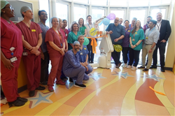 UHBW colleagues from the Children's Epilepsy Surgery Service gather to reveal the name of the paediatric neurosurgery robot alongside Gavin May from Renishaw. The team are gathered in a semi-circle in front of a window with the white and orange robot, which looks like a robotic arm, in the middle