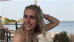 A young blonde woman is turned to her left and smiling at the camera. She is sat in an outdoor seating area of a restaurant and the sea is behind her.
