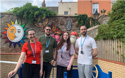 Four people (two women and two men) are standing together in a courtyard. They are all smiling at the camera. Behind them you can see a bright sun wall decoration and some play equipment