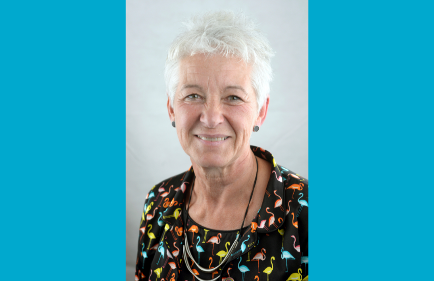 A portrait image of the new joint chair of Ingrid Barker wearing a black top with blue, red, yellow and orange flamingos against a blue background