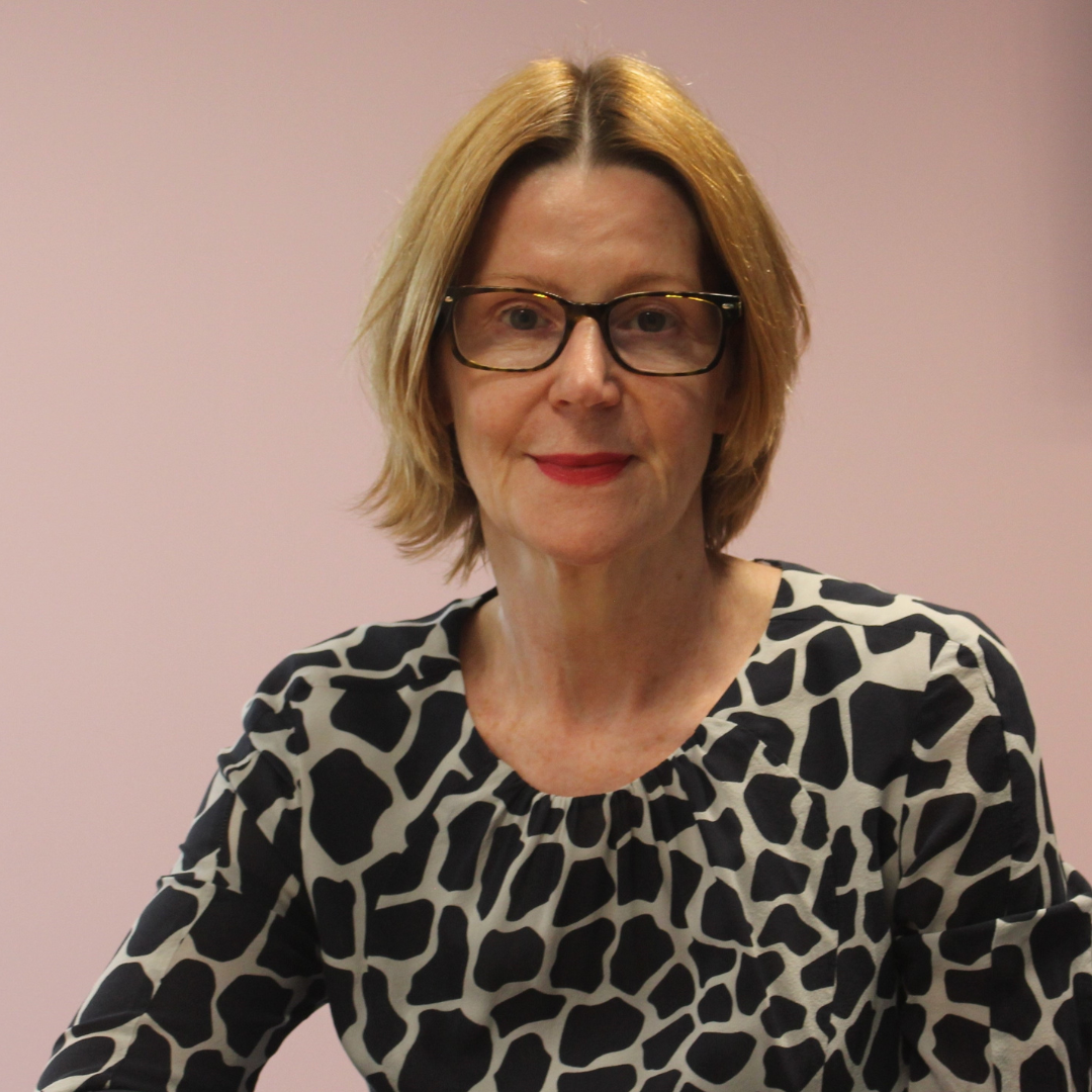 Joint Chief Executive Maria Kane has blonde hair and wears glasses and is sitting in front of a dusky pink coloured wall. Her long-sleeved top has a black and grey block pattern on it. 