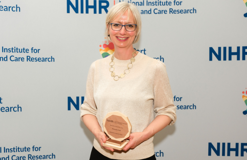 Dr Amanda Hall is holding her trophy at the awards ceremony. She is standing in front of a screen which has the National Institute for Health and Care Research logo on.