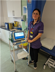 Nurse Sally Tilden is stood in her purple nurse's tunic to the left of the photo. Behind her is a hospital bed and a blue curtain. She is smiling at the camera and resting her hands on a small white trolley table on wheels. On top of the trolley is a digital screen about the size of a tablet computer. 