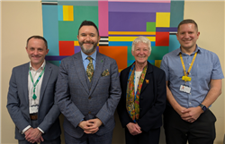 Jon, Sadik, Ingrid and Matt standing in a line in front of a piece of colourful artwork, all with their hands clasped in front of them and smiling. 