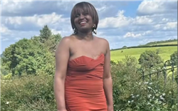 Kimi is smiling and looking to someone behind the camera. She is wearing a dark orang strapless dress which shows her mastectomy scar on the left. Behind her are trees and fields. It is a sunny day with white clouds in a blue sky. 